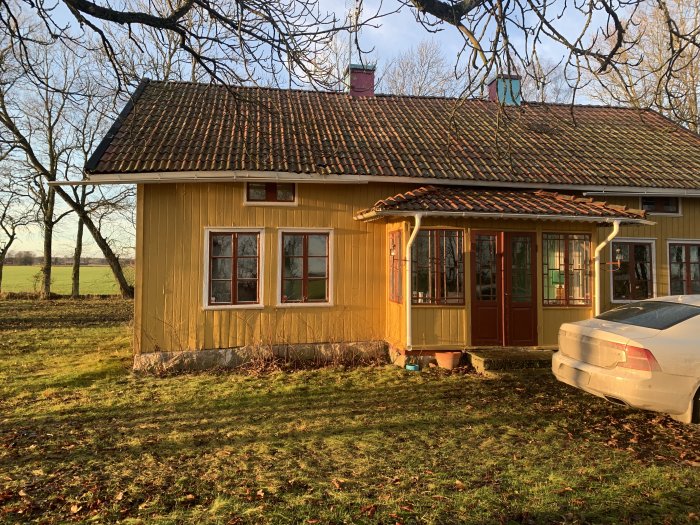 Gul träfasad på hus med röda fönster och dörr, bil parkerad framför, banmadrass och terrass.