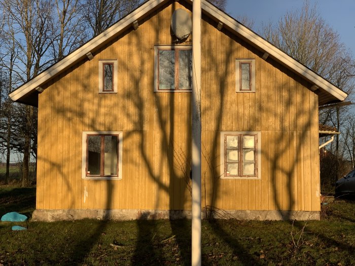 Träfasad av ett hus i solljus med olika fönstertyper och släta, rundade paneler till vänster.