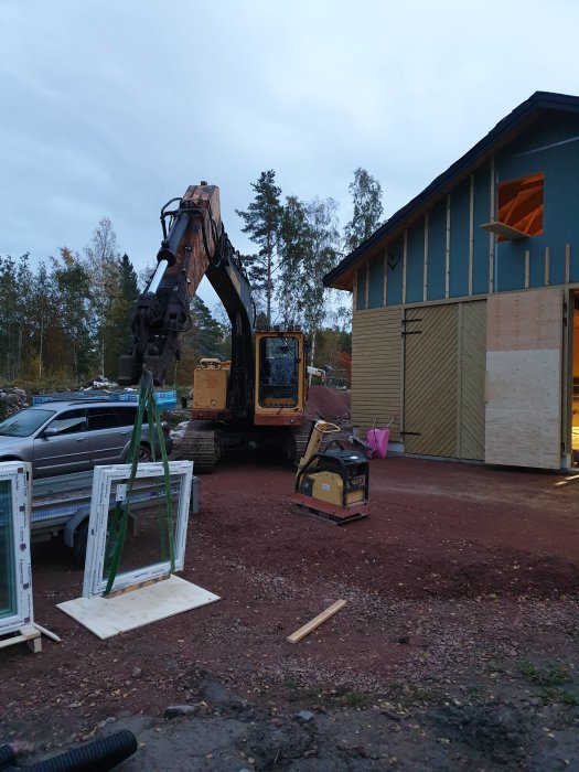 Byggplats med grävmaskin som lyfter pall med fönster, omonterade fönster och verktyg i förgrunden.