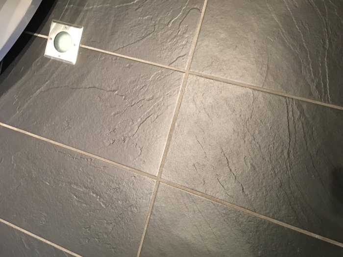 Mörkgrå klinkergolv med vita fogar och synliga kalkfläckar i ett duschutrymme.