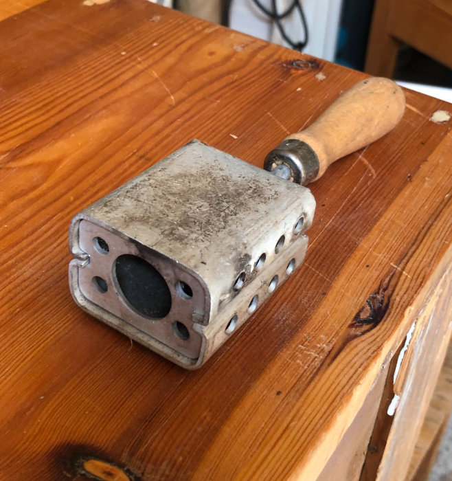 Gammalt verktyg med träskaft och metallhuvud med hål, ligger på ett träbord.