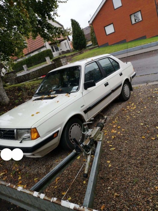 Vit Volvo 360 parkerad på en grusuppfart med höstlöv, bredvid ett hus och en släpvagn.