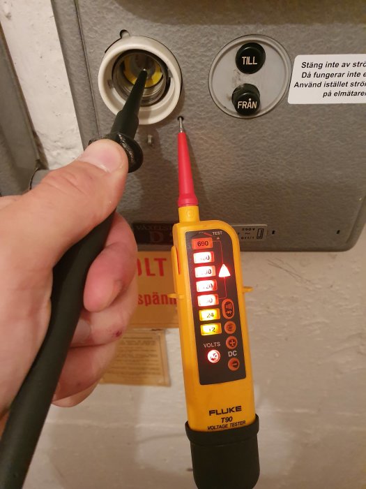 Hand som håller en spänningsprovare mot en elcentral, med en tänd lampa indikerande ström.