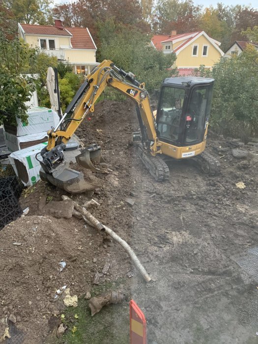 Grävmaskin på en rörig tomt under dräneringsarbete, med synligt hus och trädgård.