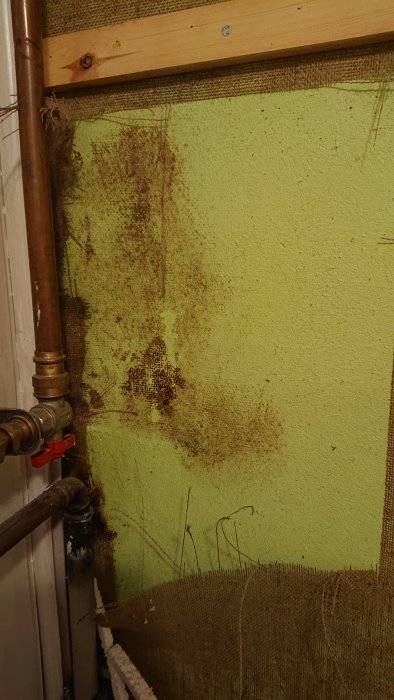 Vägg med borttagen juteväv som visar mögelskador och en röd ventil på en vattenledning.