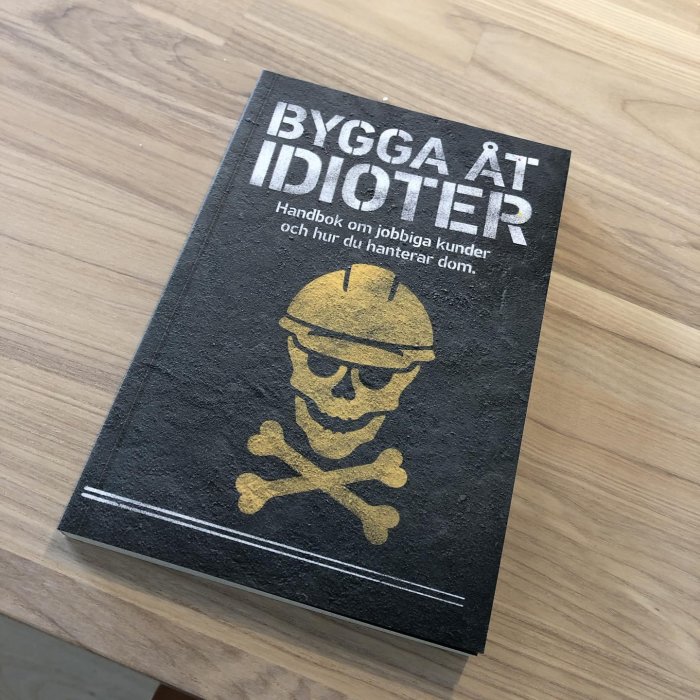 Bok med titeln "BYGGA ÅT IDIOTER" och undertext "Handbok om jobbiga kunder och hur du hanterar dom" på träbord.