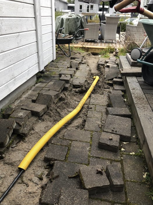 Grävd gång med gult skyddsrör bland upptagna stenplattor vid husgrund, renoveringsarbete pågår.