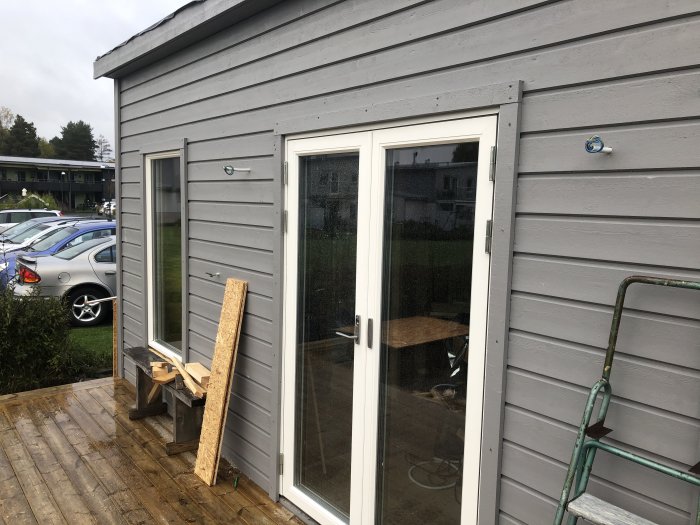 Grå träfasad med glasdörr, avsatser för bastudörrens installation, byggmaterial och en stege framför huset.