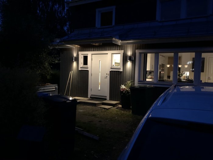 Husfasad vid skymning med nyinstallerade LED-lampor under entrétaket och öppen dörr med ljus inifrån.