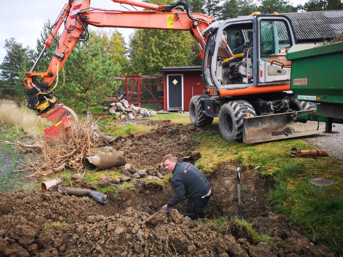 Grävmaskin utökar parkeringsyta och man reparerar avloppsrör; skavd gräsmatta och rör i förgrunden.