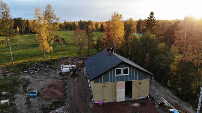 Nybyggd husgrund med pålagt svart betongpannetak omgiven av grönskande landskap och en byggarbetsplats.