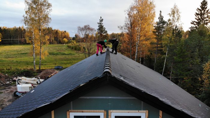 Personer lägger betongpannor på taket till en byggnad med naturen i bakgrunden.