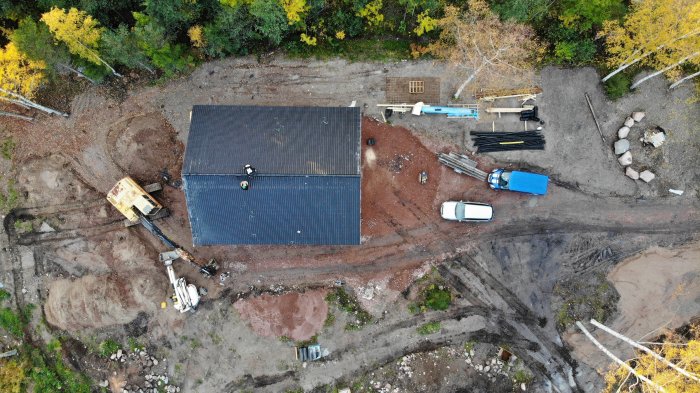 Flygbild över nybyggt hus med färdiglagt tak och material på marken, omgivet av skog och byggutrustning.