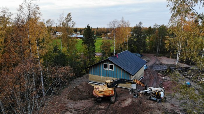 Hus med nytt tak i sluttande terräng med kranbil som lyfter byggmaterial, kringgärdad av trädbeklädd natur.