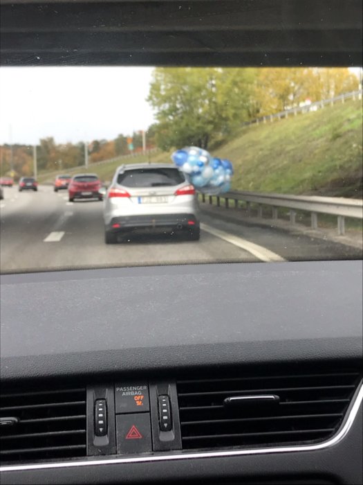 Bild från bilens insida med utsikt över väg där en annan bil har flera blå och vita ballonger bundna vid bakluckan.
