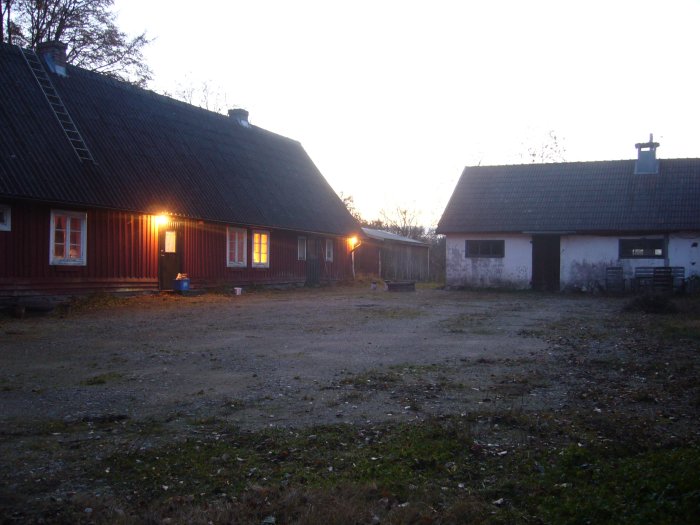 Gård med packad jord och sporadisk växtlighet, solnedgång, traditionellt rött hus och ladugård.