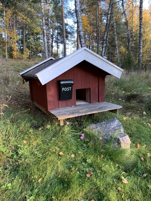 Isolerad kattkoja i trädgården med "POST" skylt på dörren, omgiven av gräs och träd.