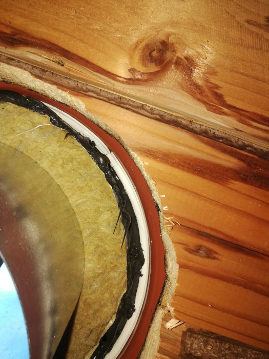 Tätning med svart tätningsmassa runt ett cirkulärt rör i ett träkonstruktionstak med isolering synlig.