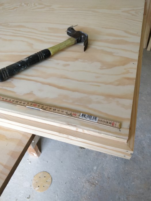 Hörn av en hemgjord plywooddörr under tillverkning med hammare och talmeter på ytan.