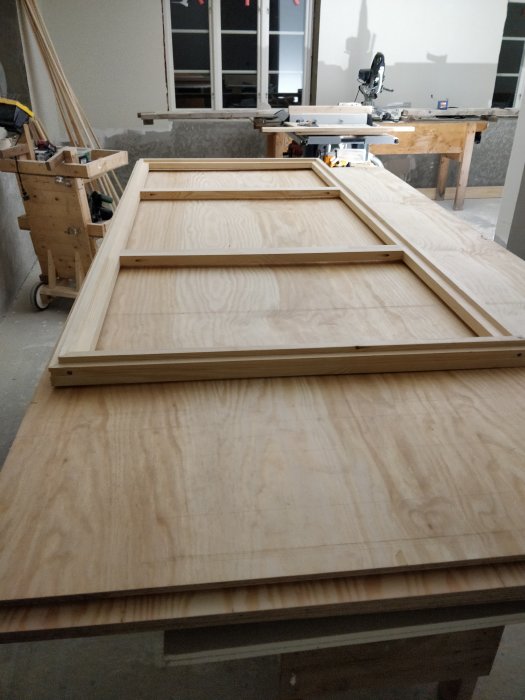 Omonterad dörrram av trä ligger på en plywoodskiva i en verkstadsmiljö med verktyg i bakgrunden.