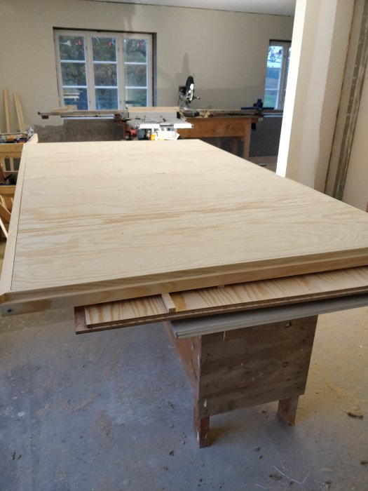 Ett nyttillverkat dörrblad av plywood ligger på ett arbetsbord i en verkstad med verktyg i bakgrunden.