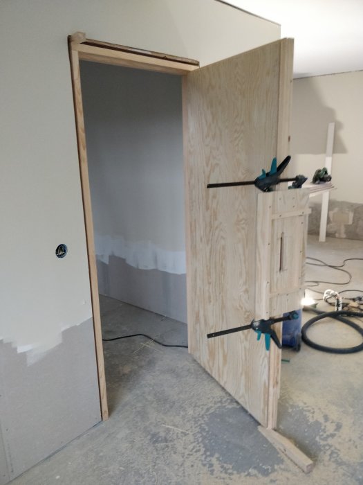 Provmonterad träddörr i öppning med klämmor, ofärdigt hål för cylinderlås synligt.