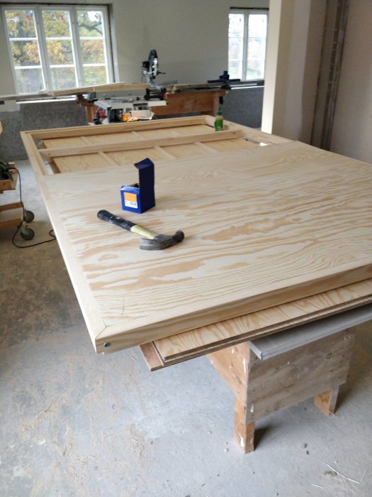 Halvfärdig träram till en dörr ligger på en arbetsbänk med plywood och verktyg i ett verkstadsrum.
