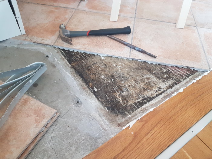 Upplyft klinkerplatta i kök avslöjar svart/brunt/rött underlag; hammare och mejsel syns.