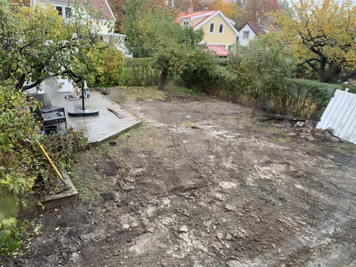 Grovåterställd trädgård med ojämnt markarbete, gräsmatta saknas med utsikt mot hus och träd.