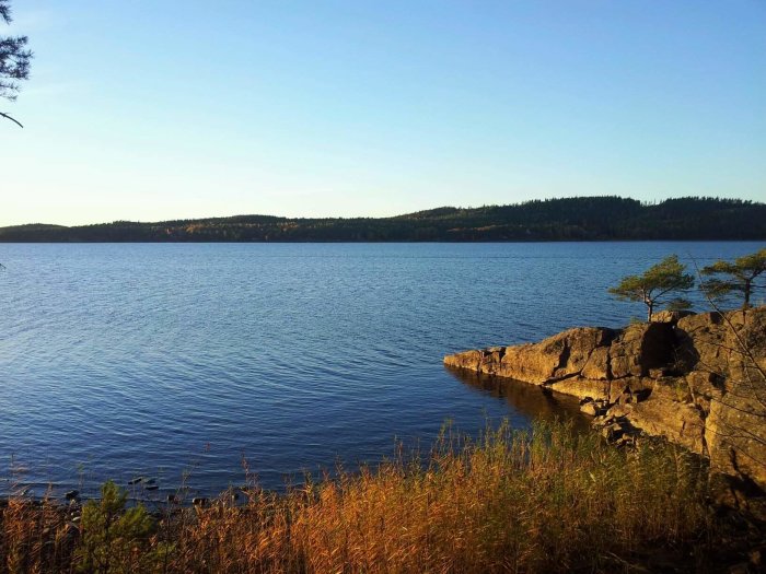 Utsikt över en sjö med klippor i förgrunden, träd och blå himmel vid solnedgång.