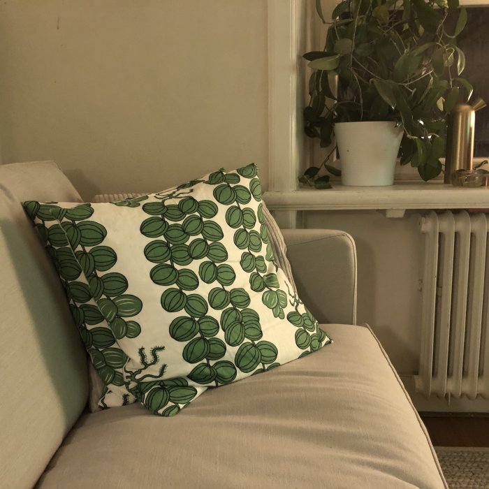 Hörnet av en ljus soffa med ett dekorativt kudde med gröna bladmönster, bredvid ett fönster med en hängande växt.