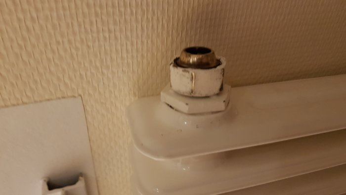 Radiatormutter med tecken på skador och repor på en vit radiator mot en tapetserad vägg.