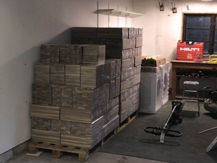 Byggmaterial staplat i ett garage med gips, plywood, och isolering nära verktygsbord och Hilti-väska.