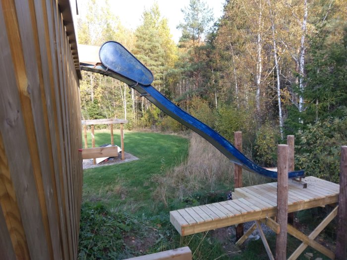 Nybyggd träplattform med en blå rutschkana och en träbro i en höstig trädgård.