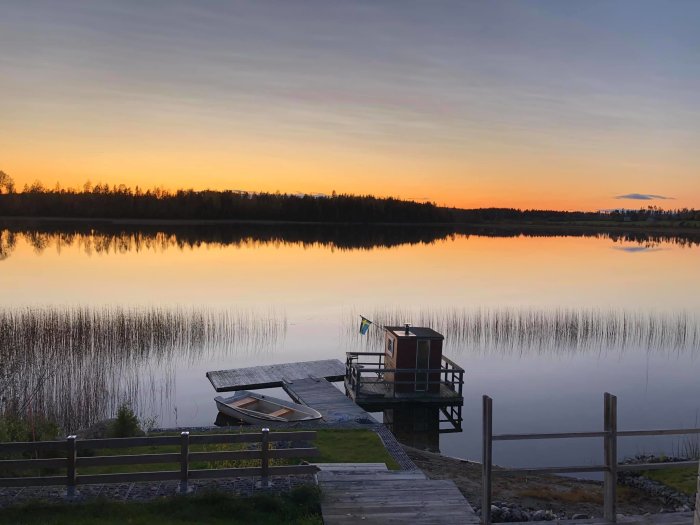 Solnedgång vid en sjö med en brygga och båt, staketet i förgrunden skulle ersättas med glasräcke.
