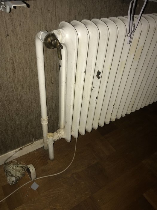 Väggmonterad radiator med avlägsnade sektioner, synliga rör och sladdar på trägolv.