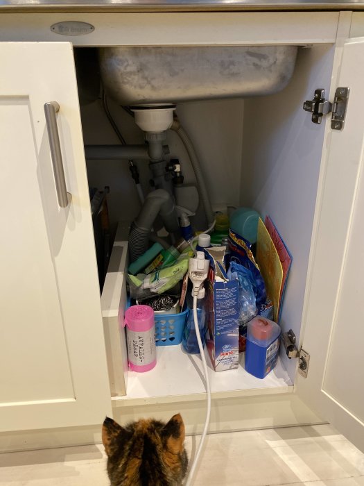 Diskmaskin kopplad till förlängningssladd under vasken, omgiven av städprodukter, med katten Smilla som tittar på.
