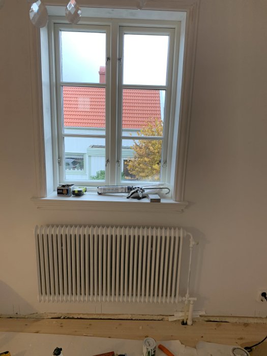 Vit radiator under ett fönster, ovanför synlig pex-slang och byggmaterial på golvet under renovering.