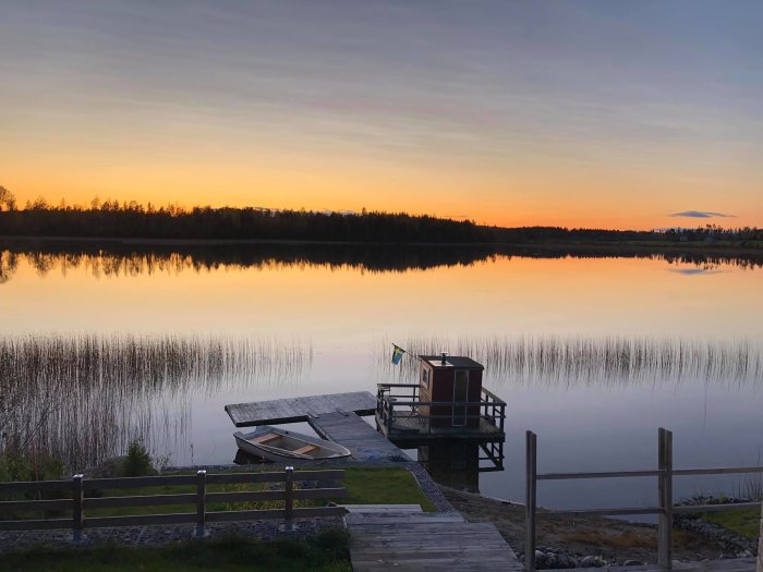 Altan vid sjö med solnedgång, bastu och roddbåt vid brygga, spegelblank vattenyta.