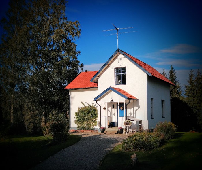 Traditionellt vitt hus med rött tak och putsad fasad, omgivet av trädgård och klarblå himmel.