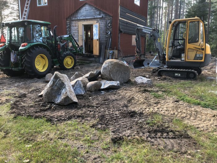 Stor sten utgrävd på gräsmatta nära röd byggnad med traktor och minigrävare i bakgrunden.