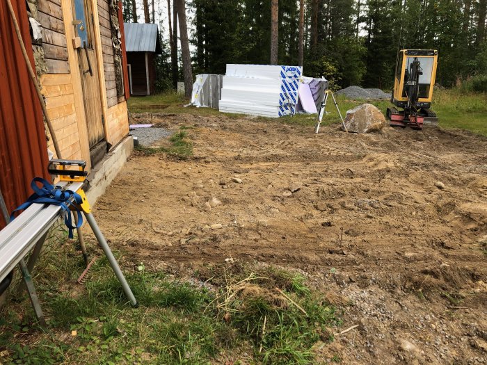Uppgrävt område nära en byggnad med en stor sten, verktyg och en minigrävare i bakgrunden.