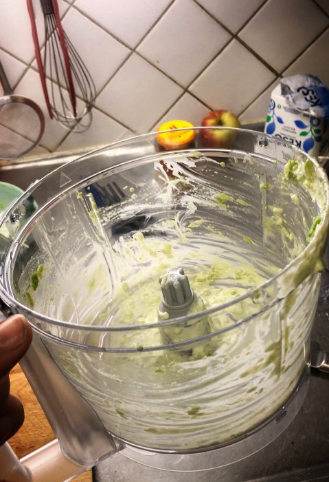 En matberedares behållare med gröna matrester på insidan, handhållen i ett kök framför köksredskap.