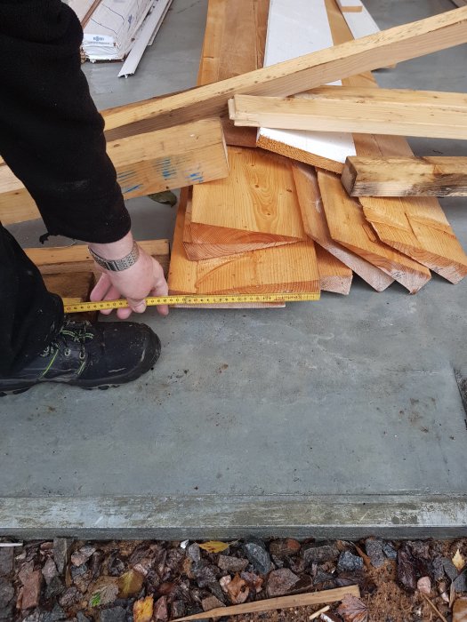 Person mäter en kluven träbräda kilformad bland andra brädor på ett betongunderlag.