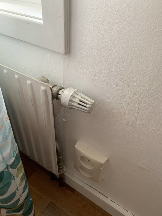 Gammal termostat med vridknopp på en vit radiator vid fönstervägg.