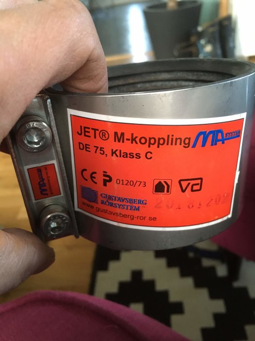En hand håller en Jet-koppling för avloppsrör märkt 75 mm, mot en inomhusbakgrund.
