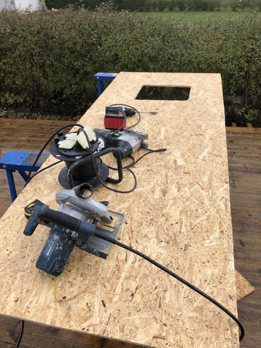 OSB-skiva med utsågat hål och verktyg som sticksåg och slipmaskin på arbetsbänk utomhus.