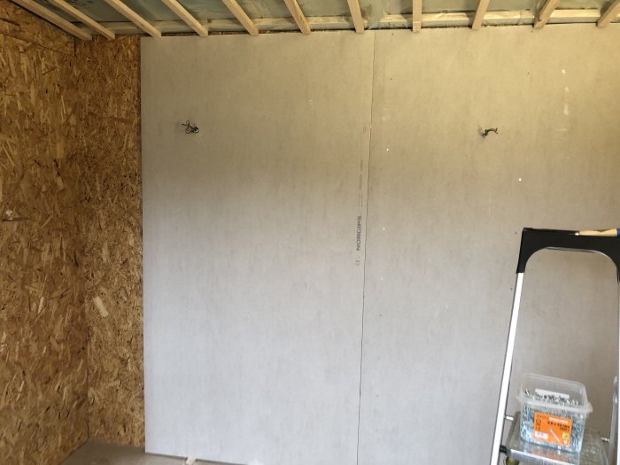 Gipsvägg under uppsättning mot en OSB-skiva med synlig isolering och trästomme, en stege till höger.