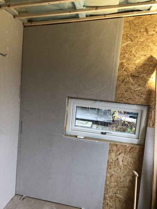 Vägg i ett rum under renovering där gipsskivor har monterats runt ett fönster, med OSB-skivor synliga på en annan vägg.