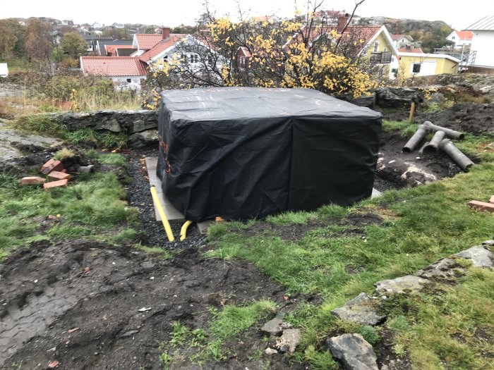 Ett spa täckt med svart presenning i en oavslutad trädgård med gräsmatta och stenläggning.